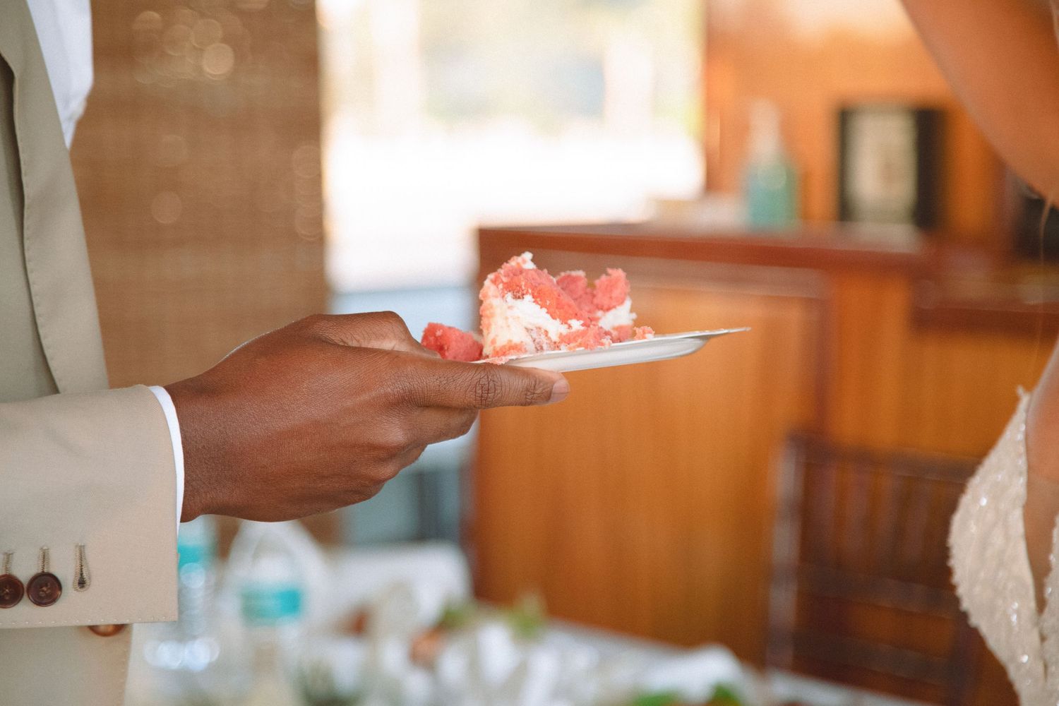 Close-up of groom holding white plate with slice of red velvet wedding cake at reception.