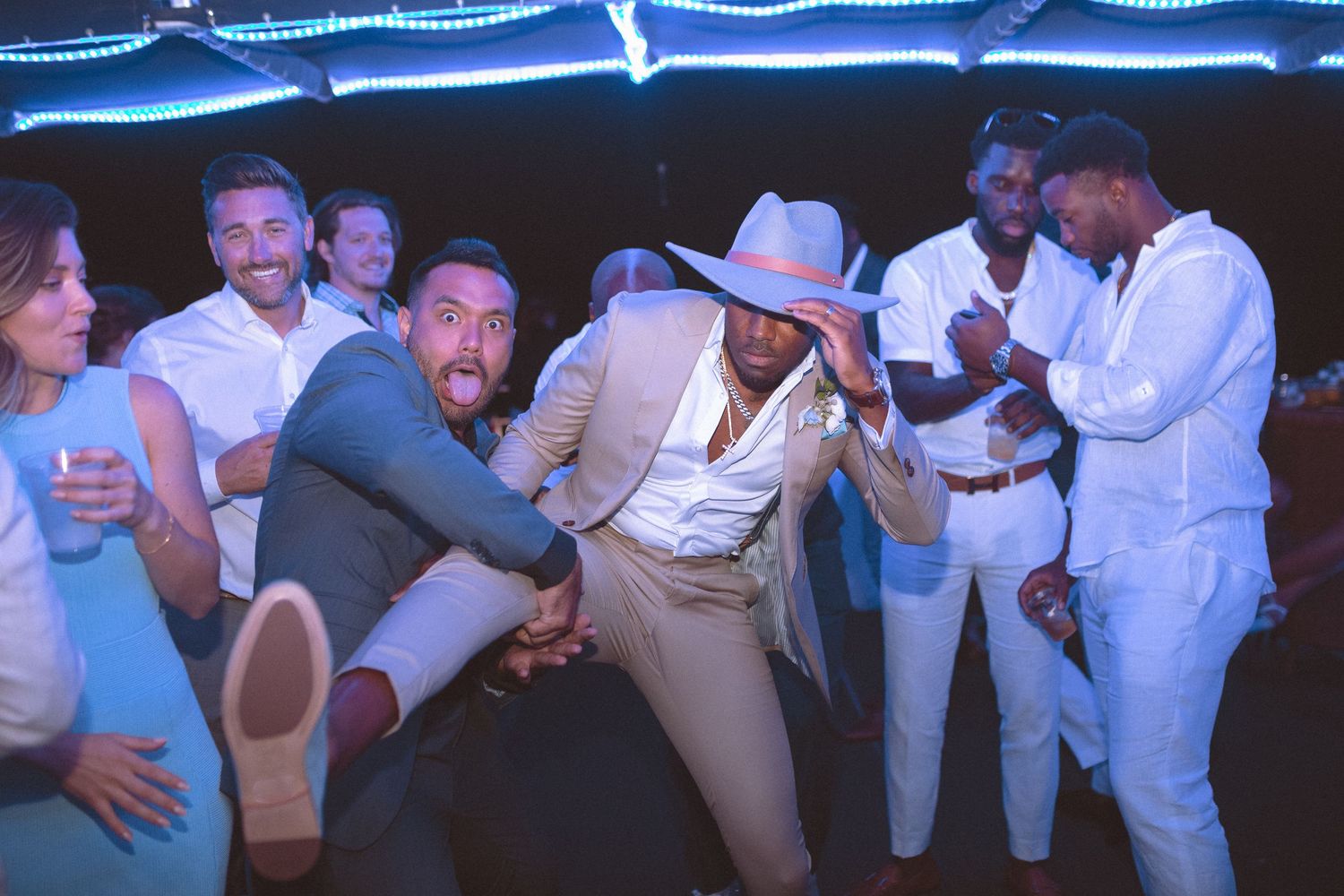 Groom in tan suit and hat dances energetically with groomsmen at lively wedding reception party.