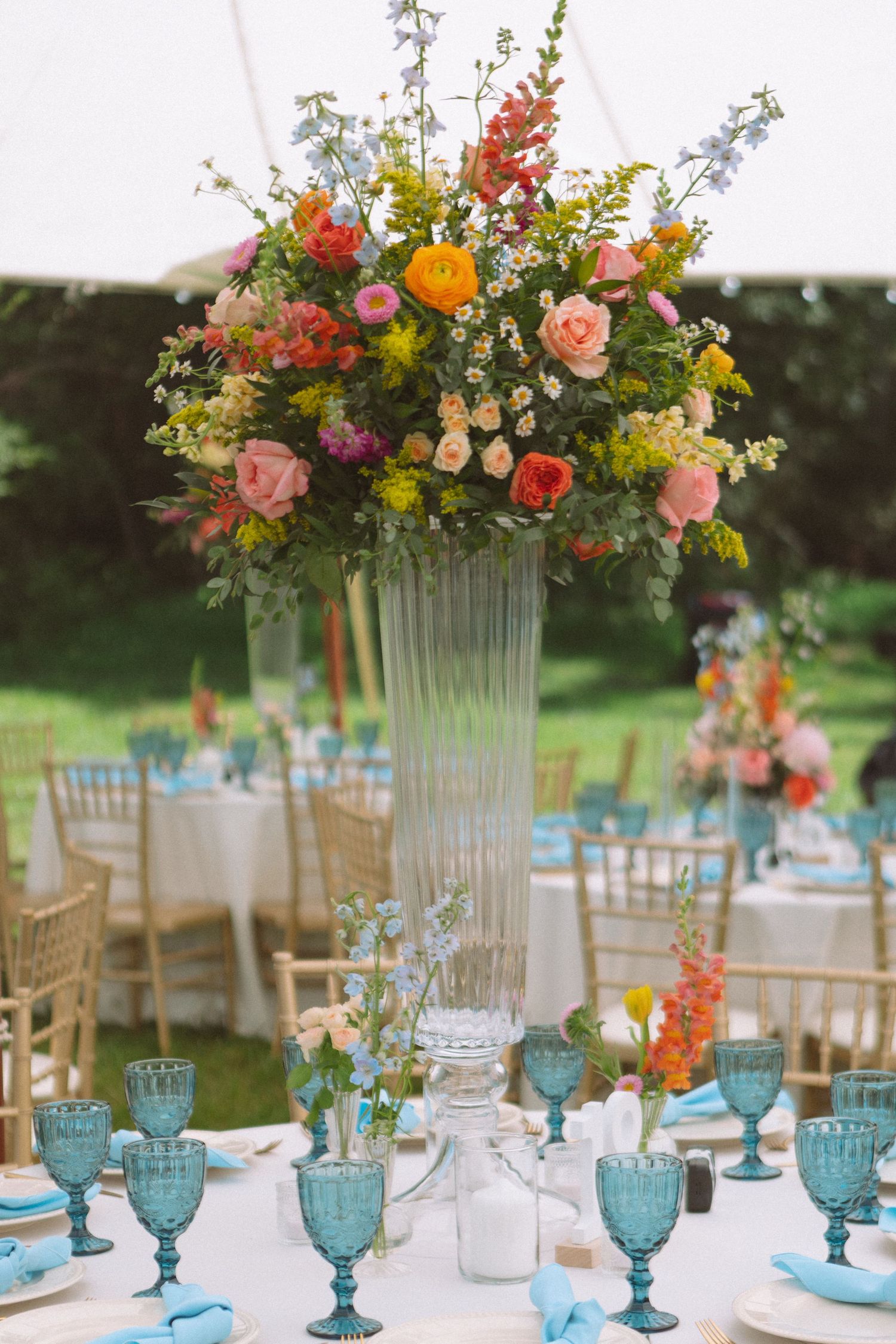 Tall glass vase with colorful floral centerpiece on wedding reception table with blue goblets.