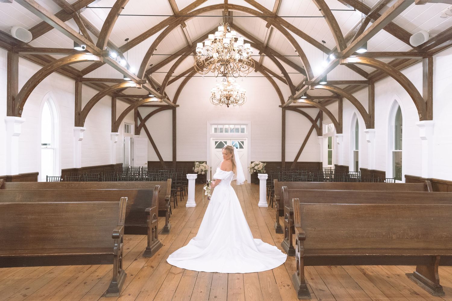 Bride standing at altar of rustic white chapel with wooden pews and arched ceiling.