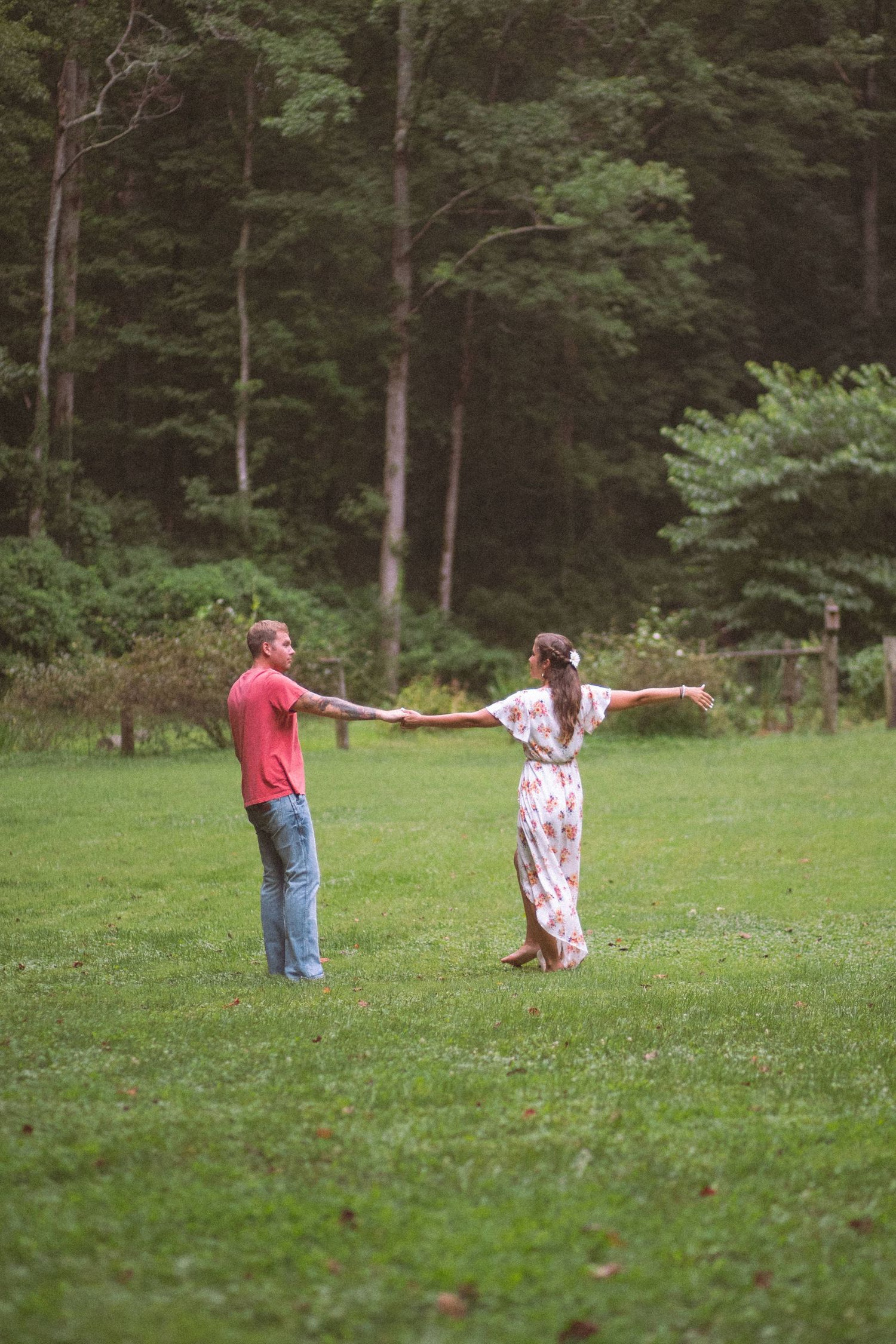Couple holding hands in a lush green meadow surrounded by tall trees, woman in floral maxi dress with arms outstretched.
