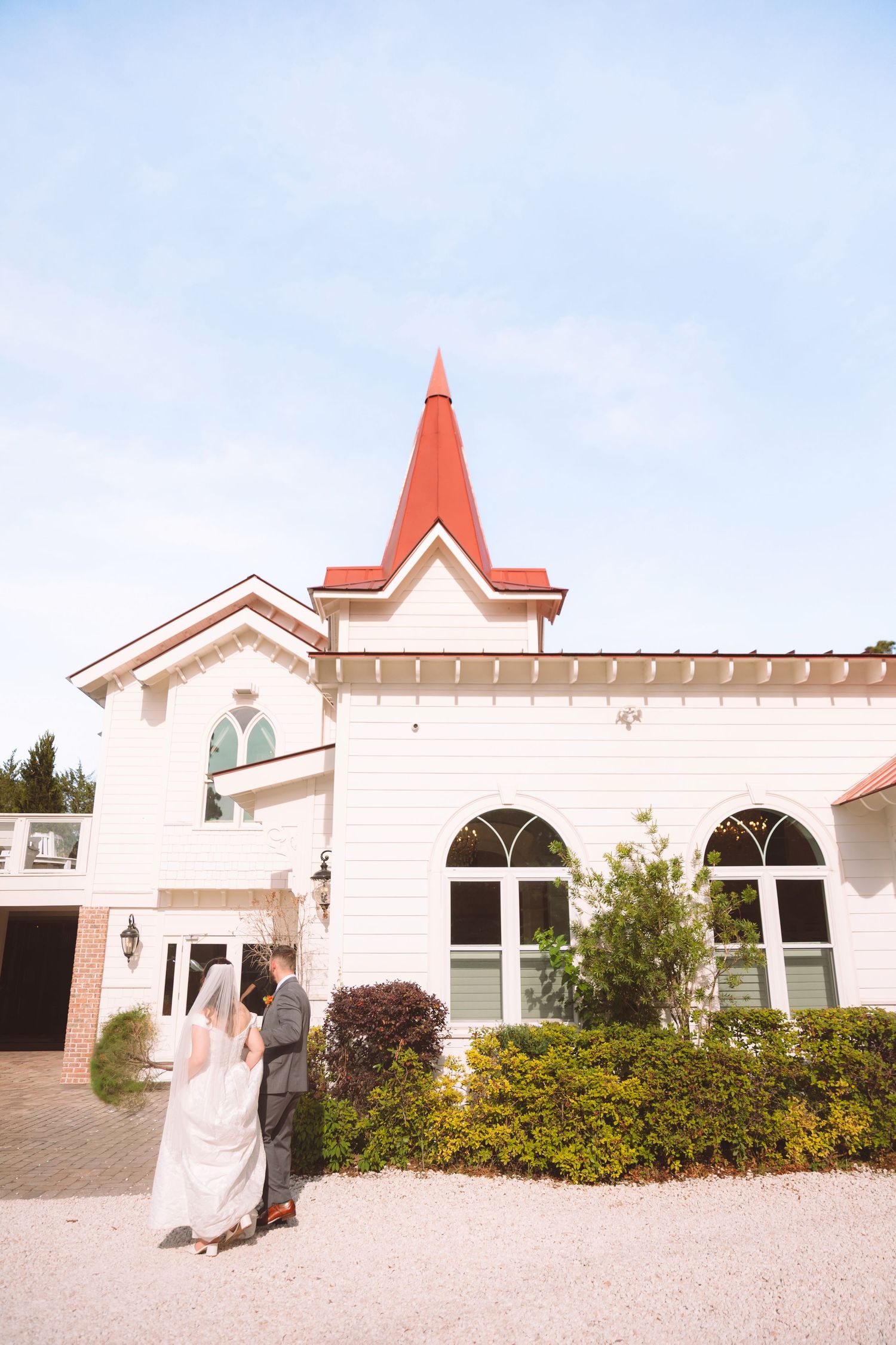Bride and groom stand before a white chapel with a red spire surrounded by lush greenery under a blue sky.