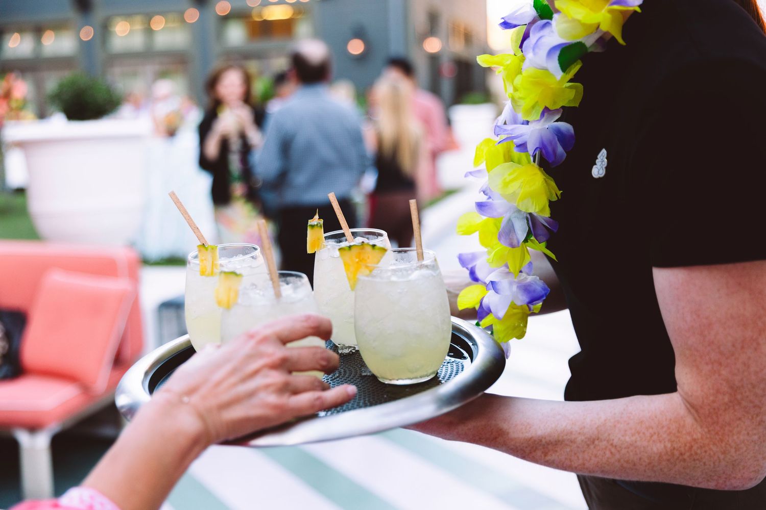 Server wearing a lei carries a tray of tropical lemonade cocktails at an outdoor summer party event.
