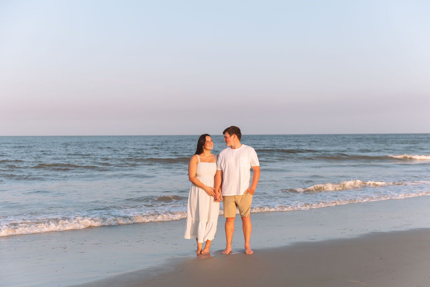 A couple holds hands on a sandy beach at sunset, wearing white outfits, with gentle ocean waves in the background.