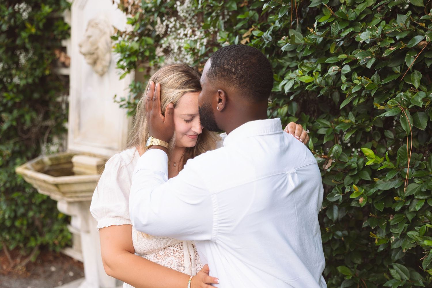 An interracial couple shares a tender moment near a garden wall with lush greenery.