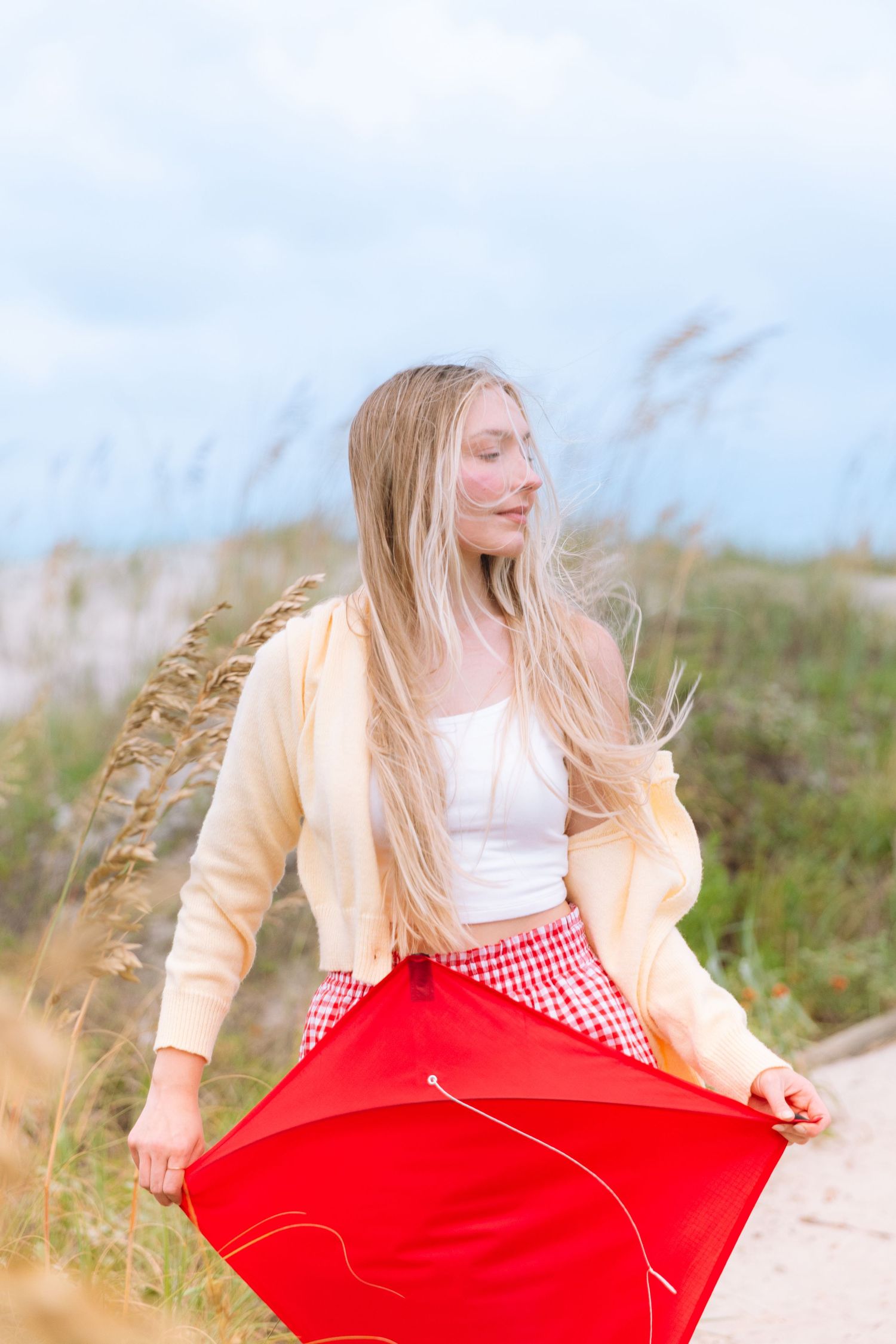 Blonde woman holding red kite on sandy beach path with sea oats in wind.