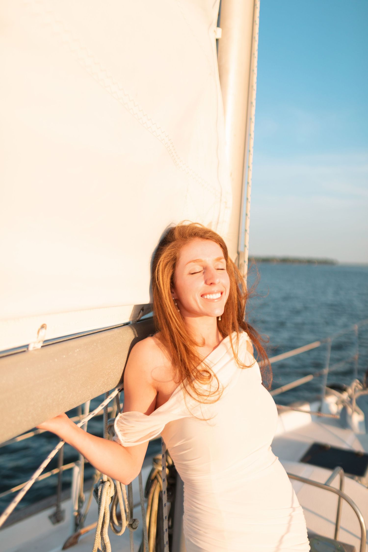 A joyful woman in a white dress smiles with eyes closed on a sailboat deck at golden hour.