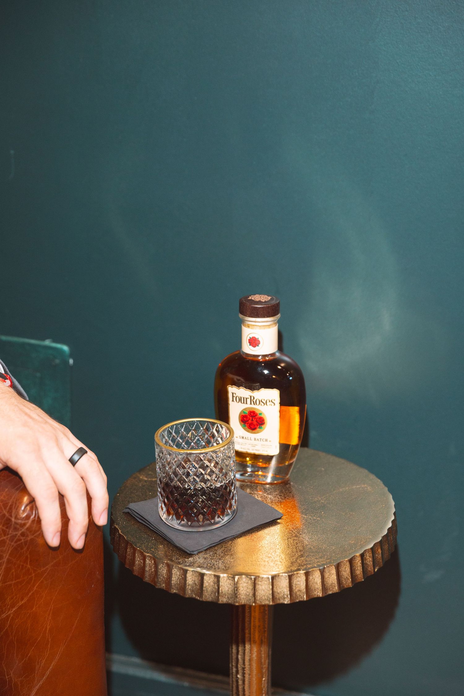 Four Roses bourbon bottle with crystal rocks glass on gold side table against teal wall.
