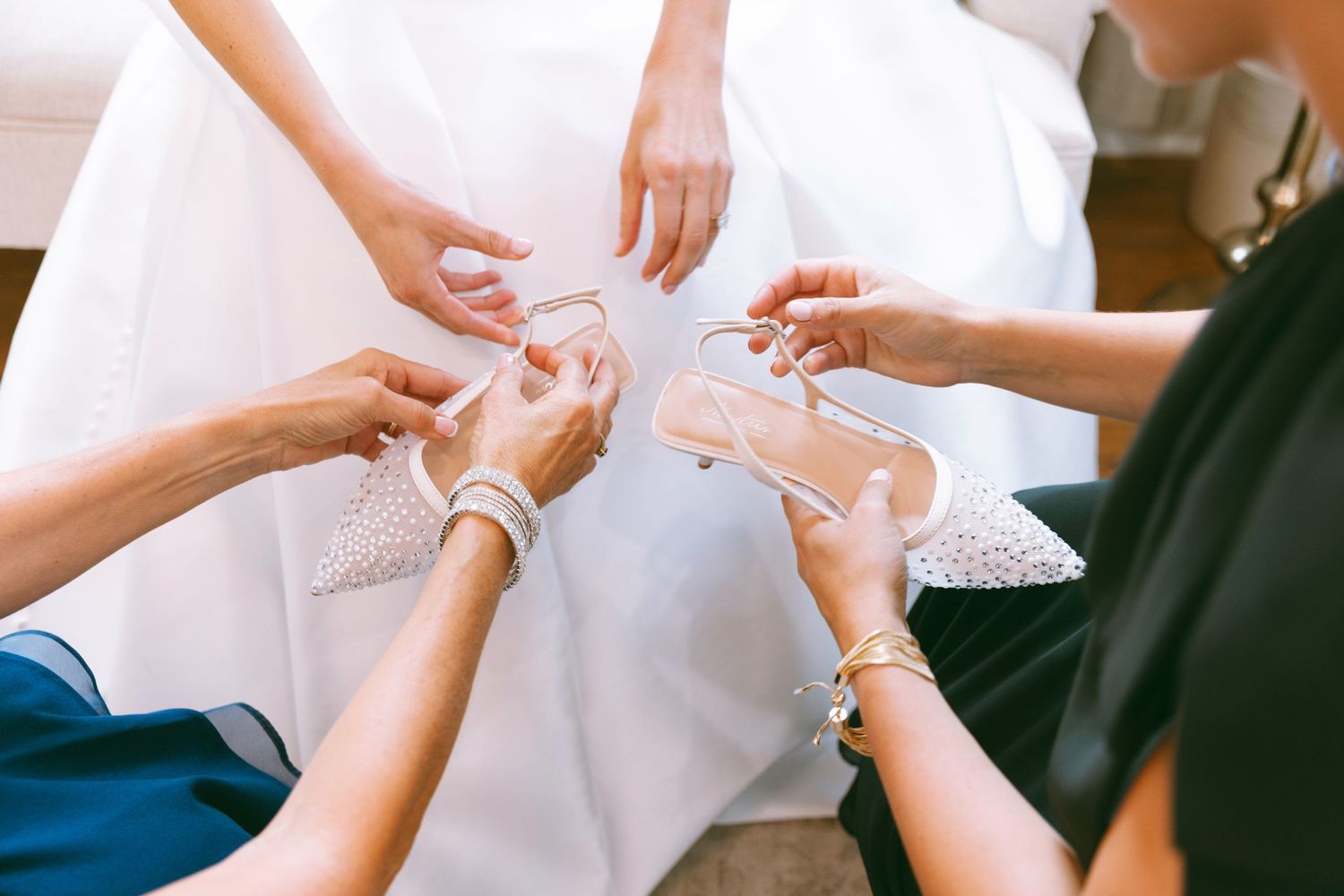 Bridesmaids holding elegant bridal heels around a bride in a white wedding dress during wedding preparations.