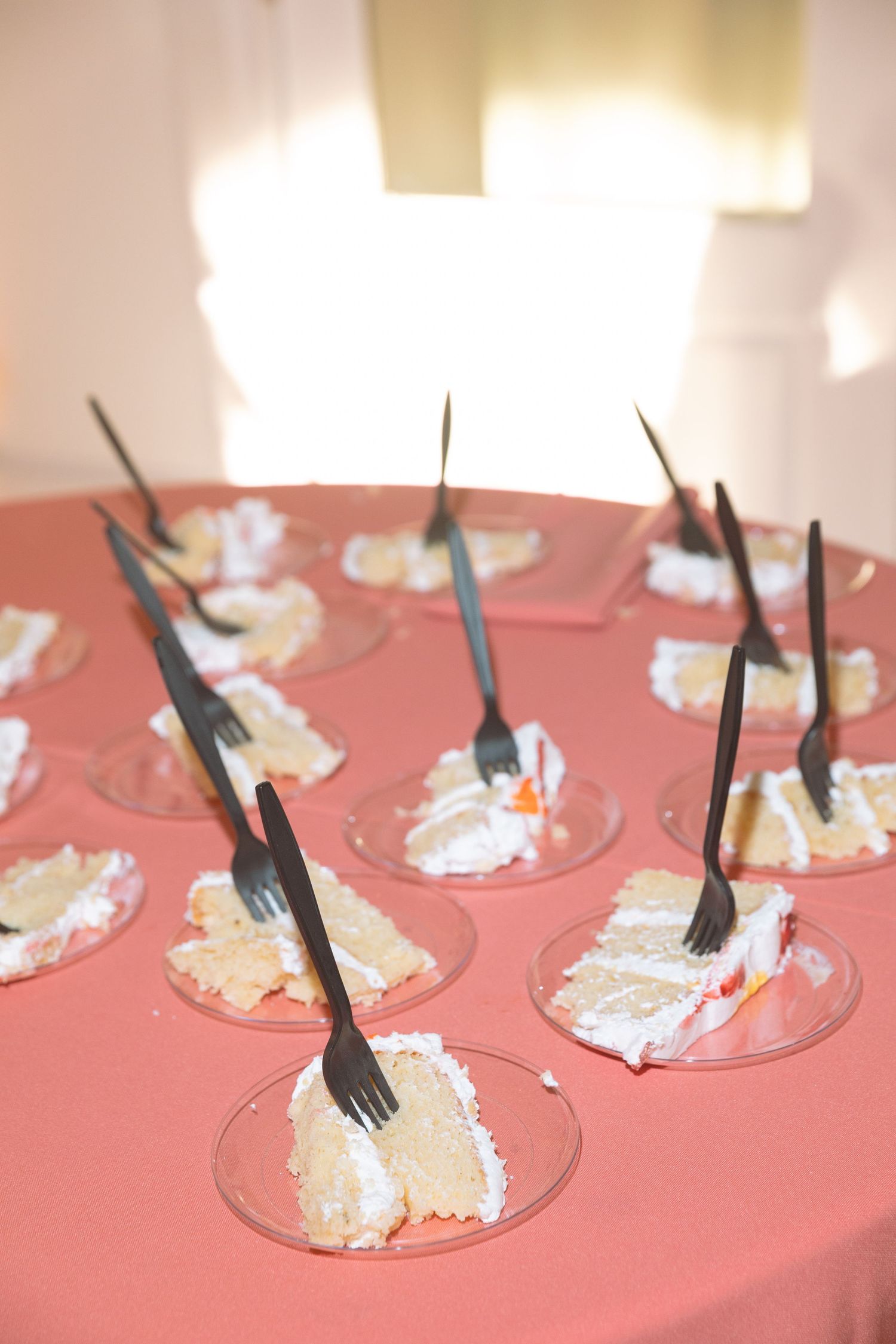 Slices of white wedding cake on clear plates with black forks, arranged on a pink tablecloth at a reception.