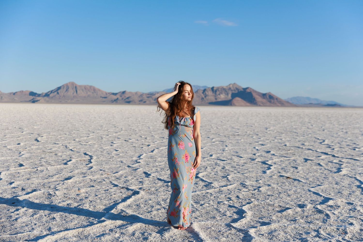Woman in floral maxi dress poses on Bonneville Salt Flats with mountain backdrop under clear blue sky.
