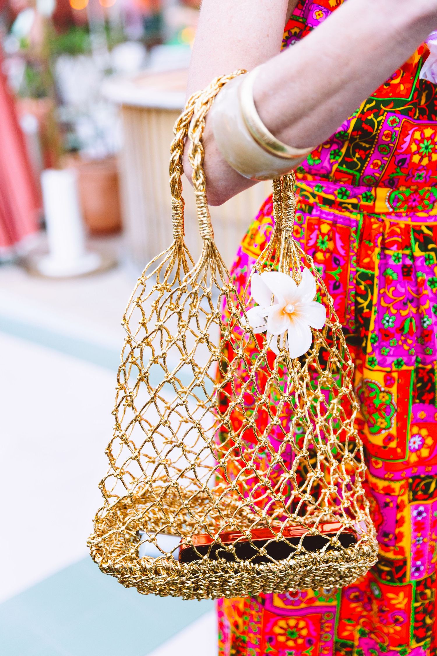 Woman holding a gold woven mesh handbag with white flower charm, wearing colorful floral dress and gold bangles.