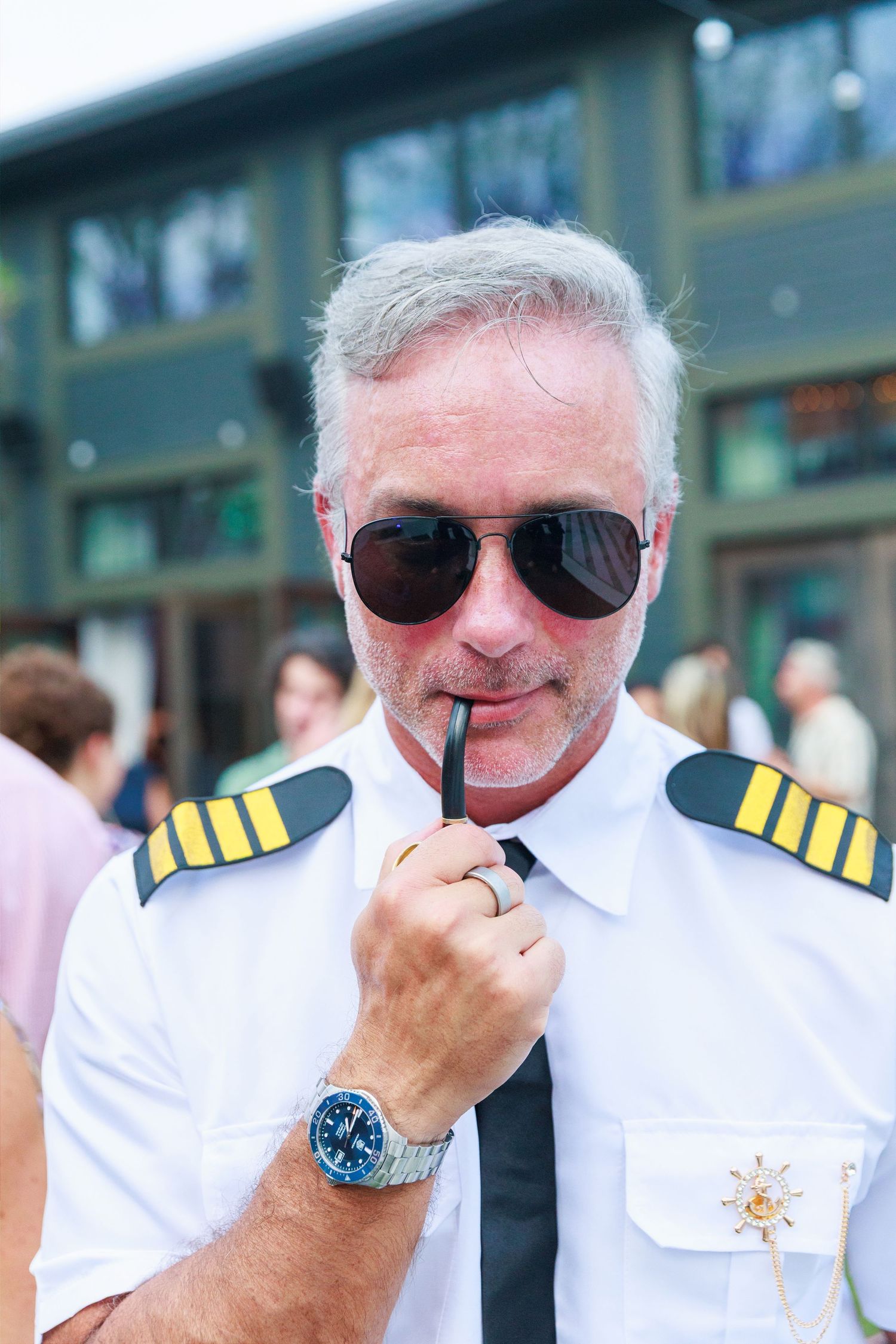 Middle-aged man in pilot uniform with epaulettes, sunglasses, and watch, holding glasses outdoors.