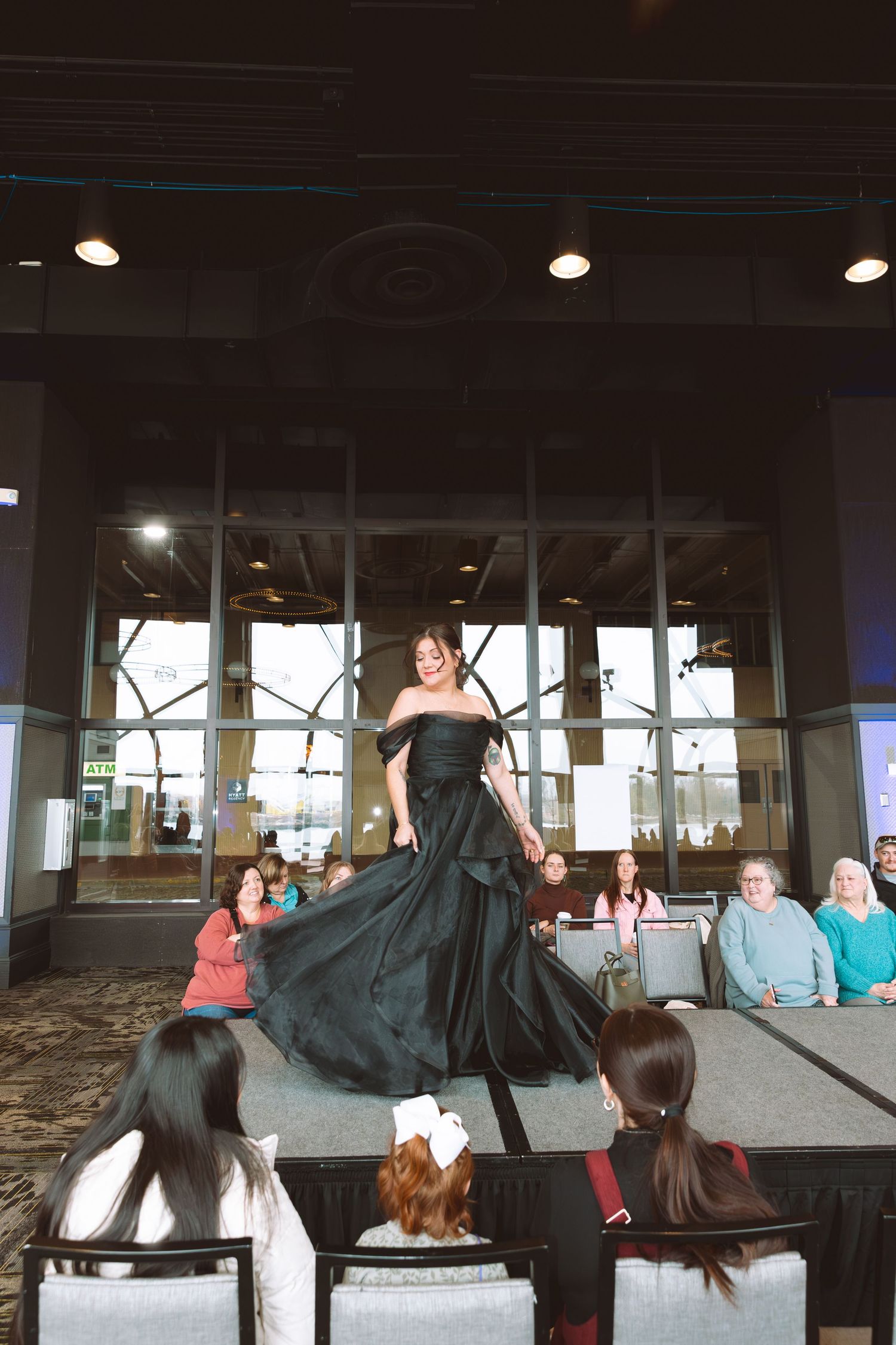 Woman in elegant black off-shoulder gown twirling on runway at indoor fashion show with seated audience.