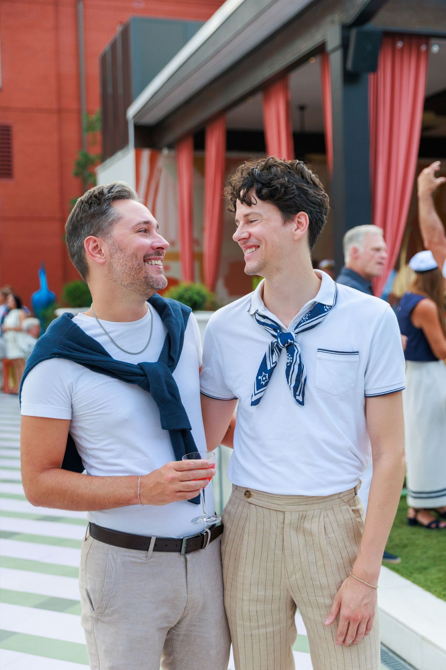 Two smiling men in white shirts with blue accessories laugh together outdoors at a social event.