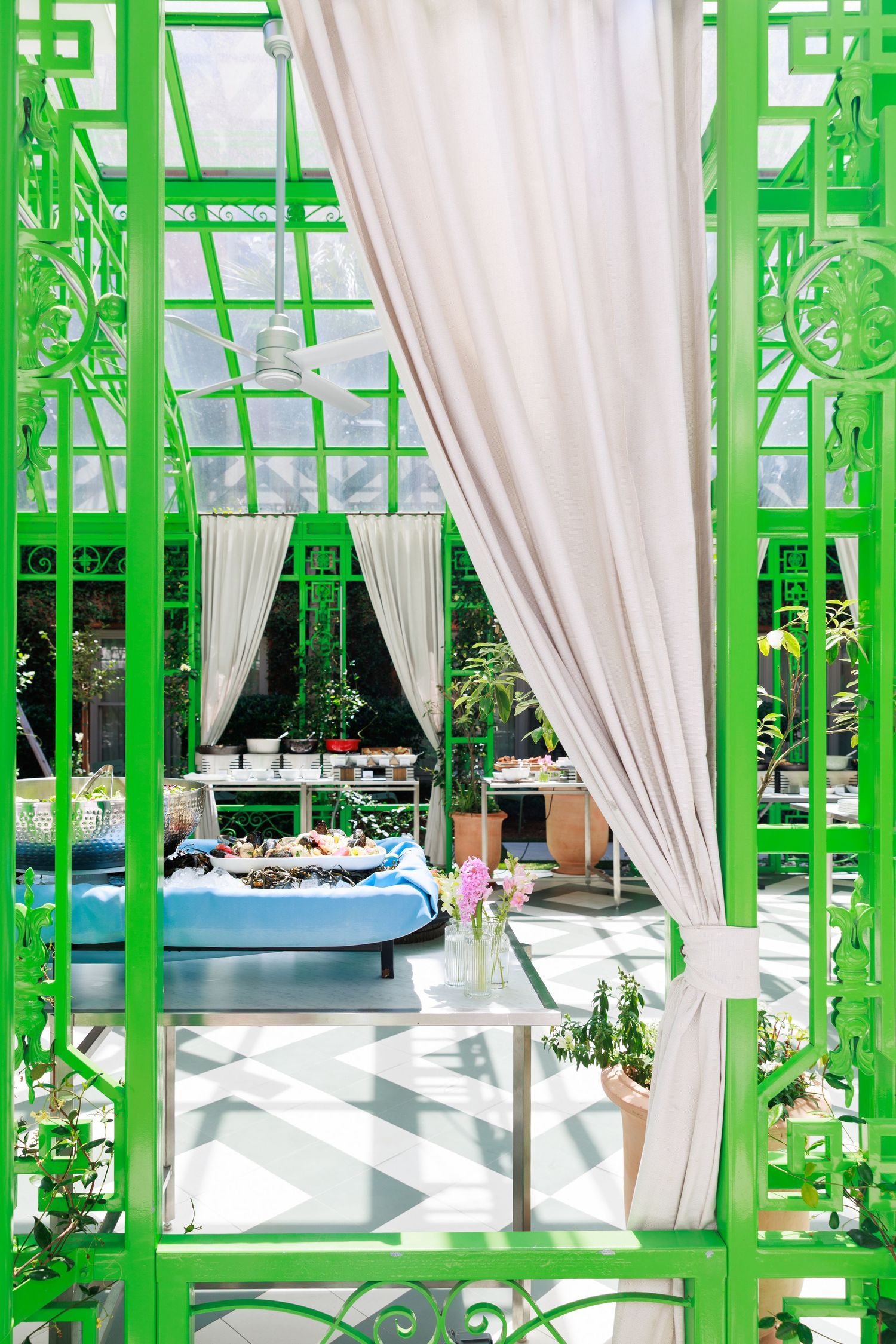 Elegant green iron gazebo with flowing white curtains, blue daybed, and lush plants in a sunlit greenhouse setting.