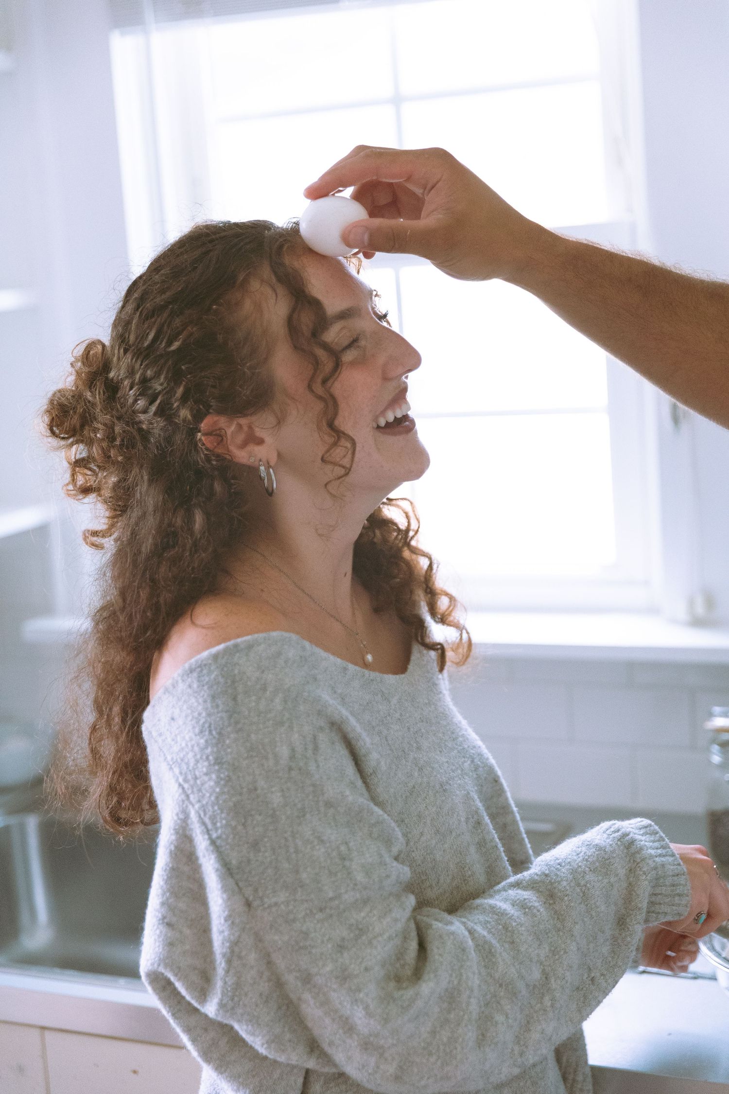 Laughing curly-haired woman in gray sweater enjoying a playful moment in a bright bathroom.