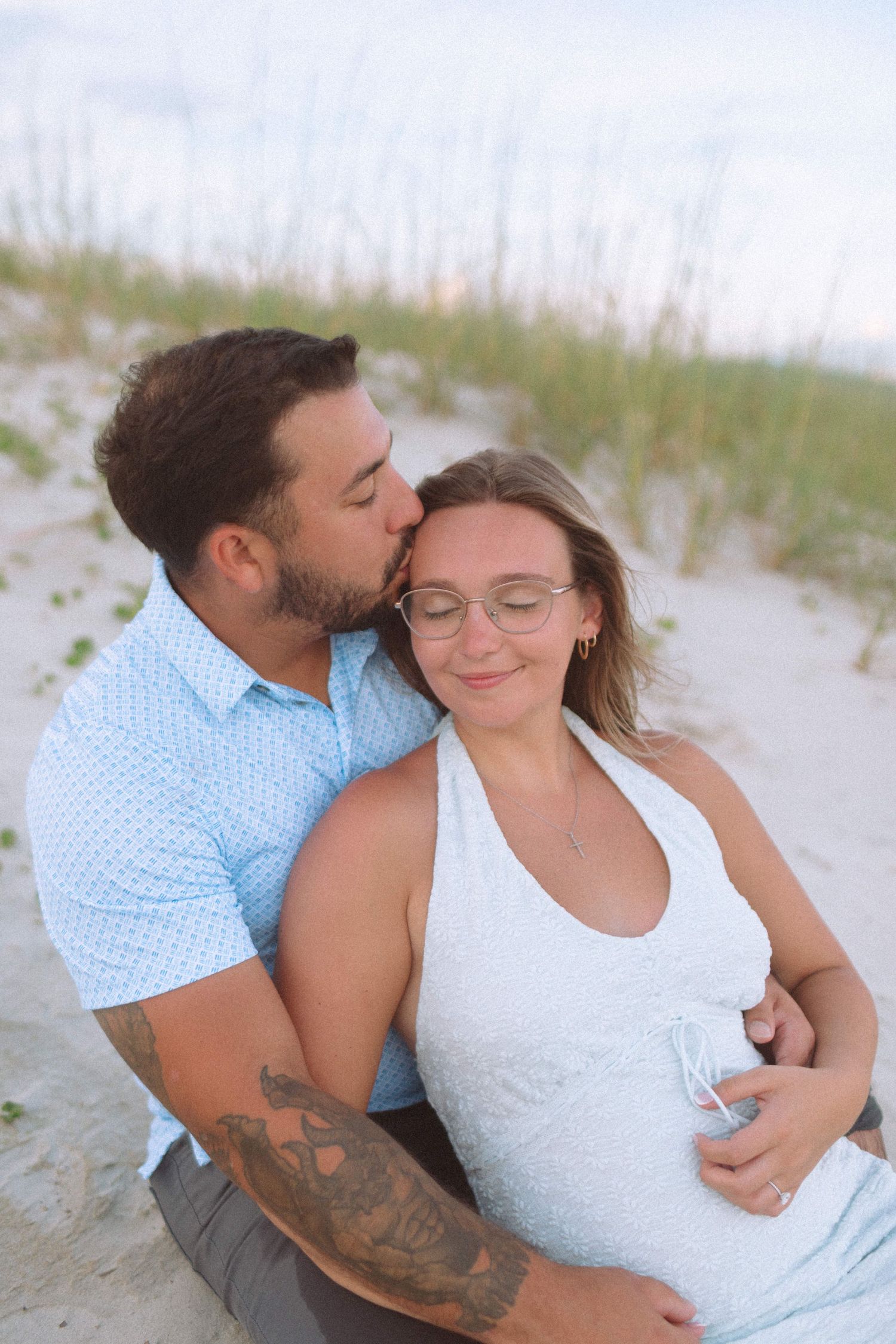 Pregnant couple sitting on beach sand dunes, man kissing womans head while embracing her bump.