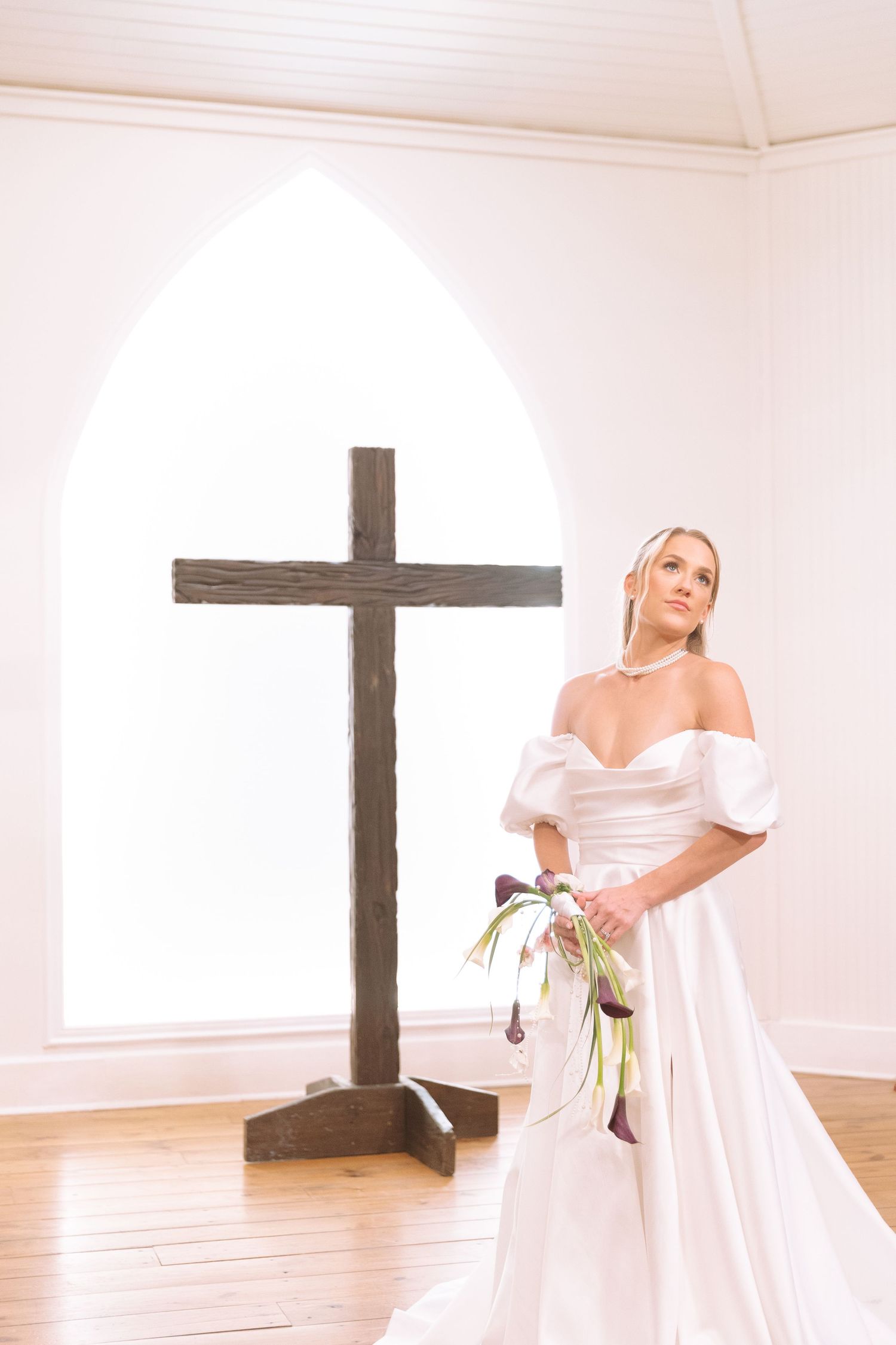Elegant bride in white satin gown standing beside a wooden cross in a bright chapel.