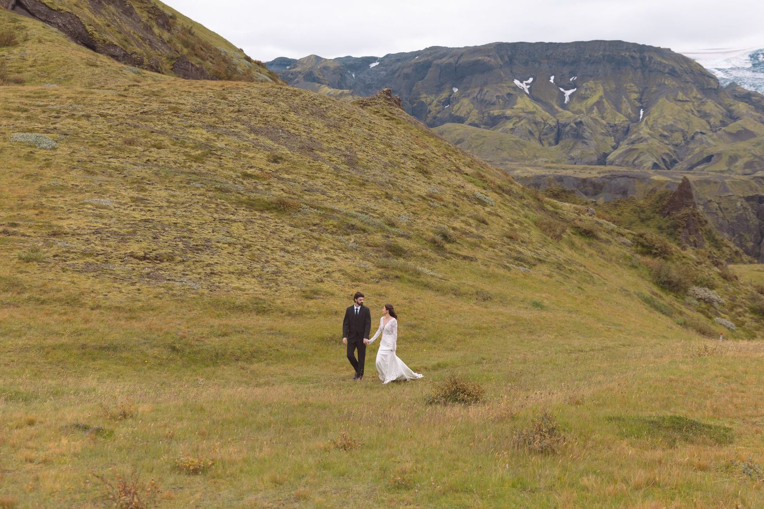 Bride and groom walking hand in hand through lush green Icelandic highlands with dramatic mountains backdrop.
