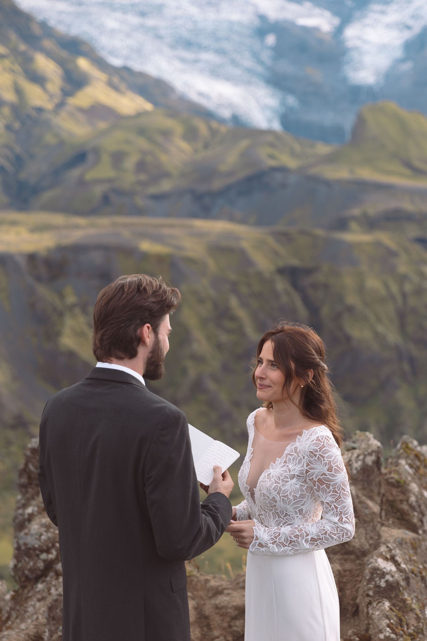 Bride smiles lovingly at groom during outdoor Iceland elopement with glacier and green mountains behind.