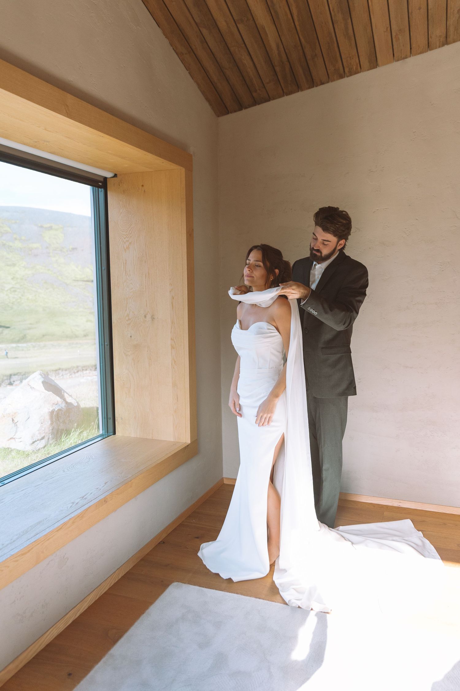 Groom adjusting brides elegant halter wedding dress near a large scenic window.