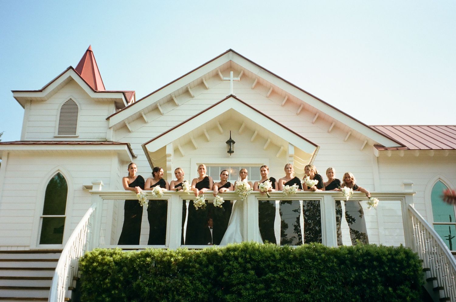 Wedding party posing on white church porch with bridesmaids in black dresses holding bouquets on a sunny day.