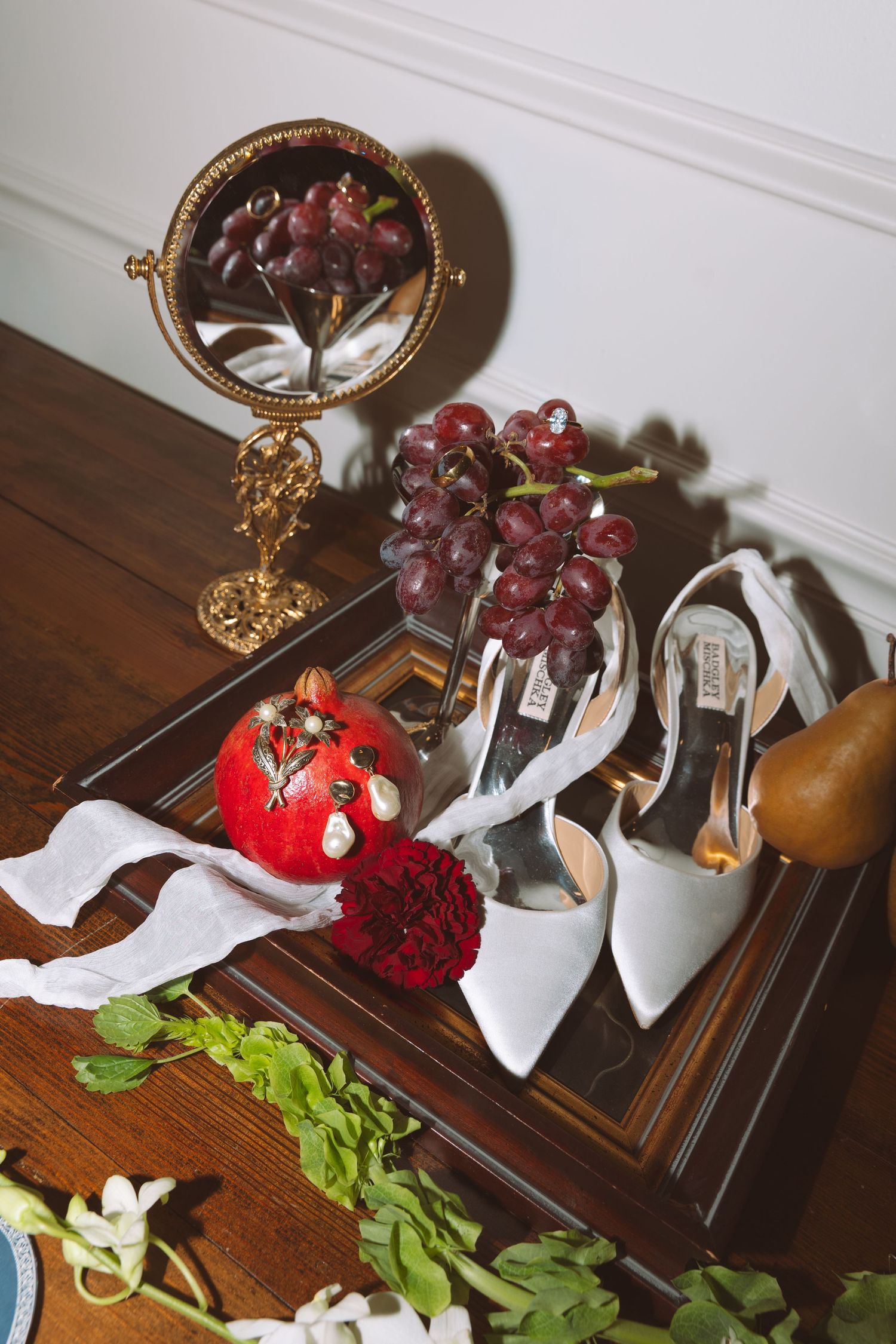 Wedding shoes displayed with grapes, pomegranate and mirror on wooden dresser.