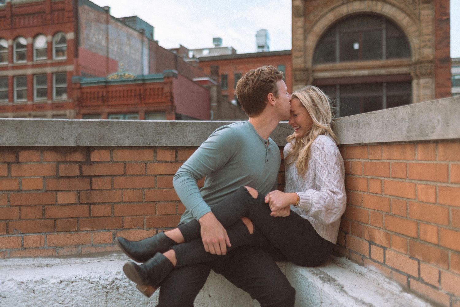 Couple sitting close together on a rooftop ledge with brick buildings behind them.