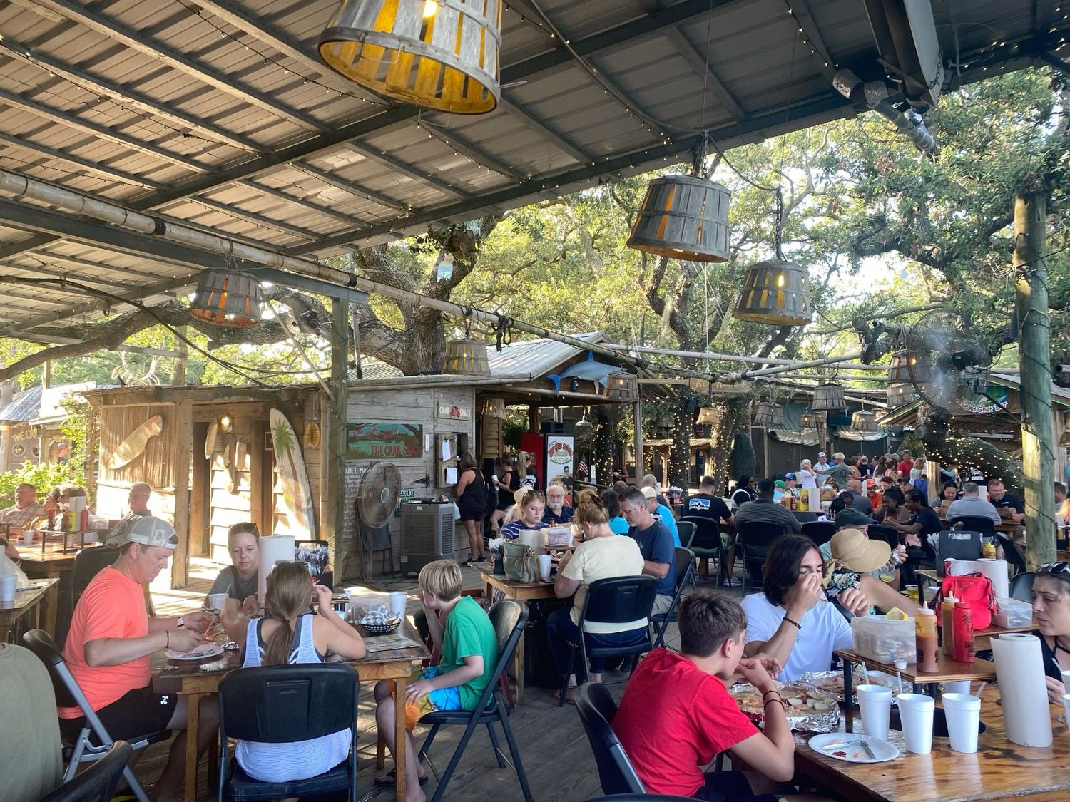 Busy outdoor restaurant dining area under rustic wooden canopy with hanging lights and large trees.
