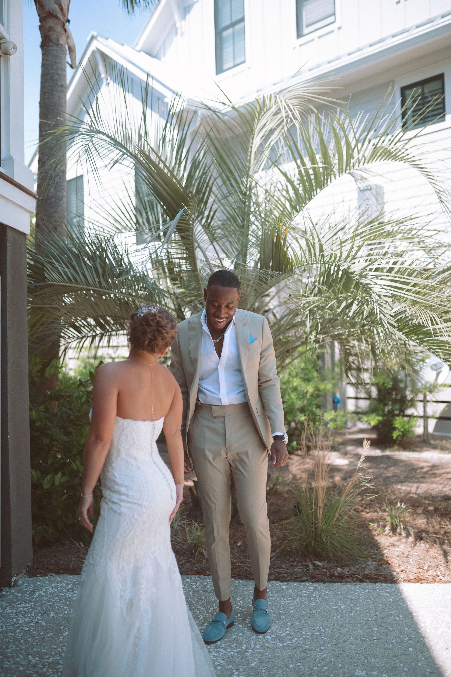 Bride in lace mermaid gown and groom in tan suit hold hands during outdoor first look near palms.