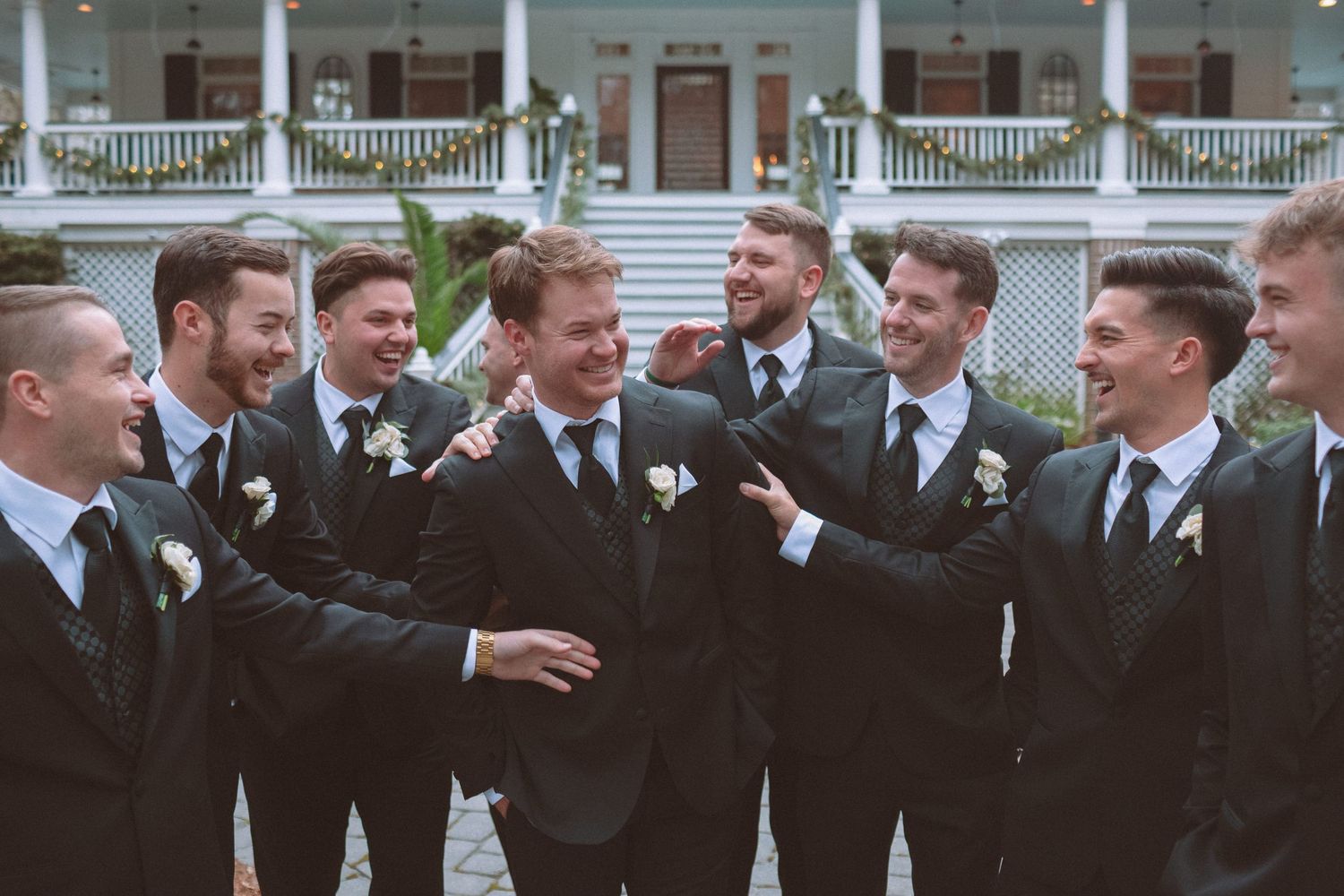 Groomsmen in black suits laugh and celebrate together outside a white colonial-style wedding venue.