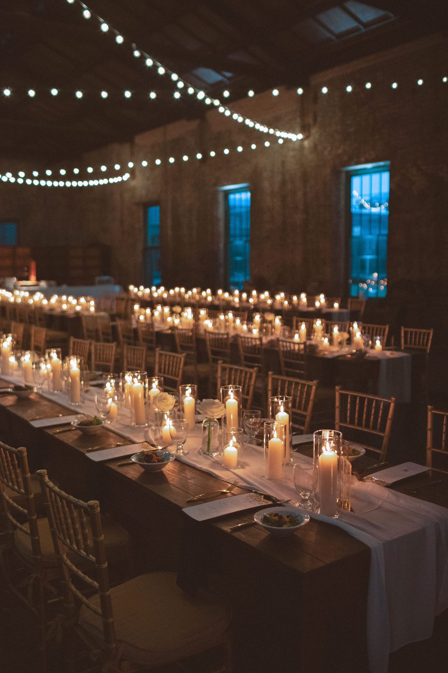 Elegant wedding reception hall with long candlelit tables, chiavari chairs, and string lights in a brick venue.