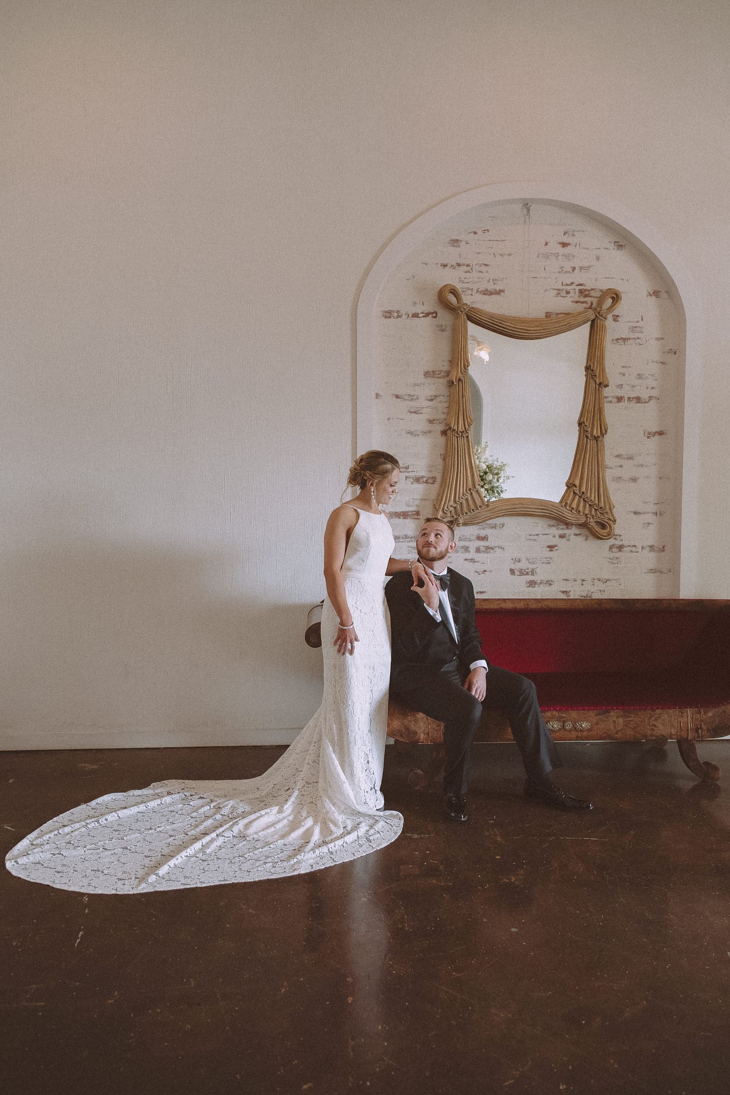 Bride in sleek white gown with lace train poses with seated groom near arched brick alcove.