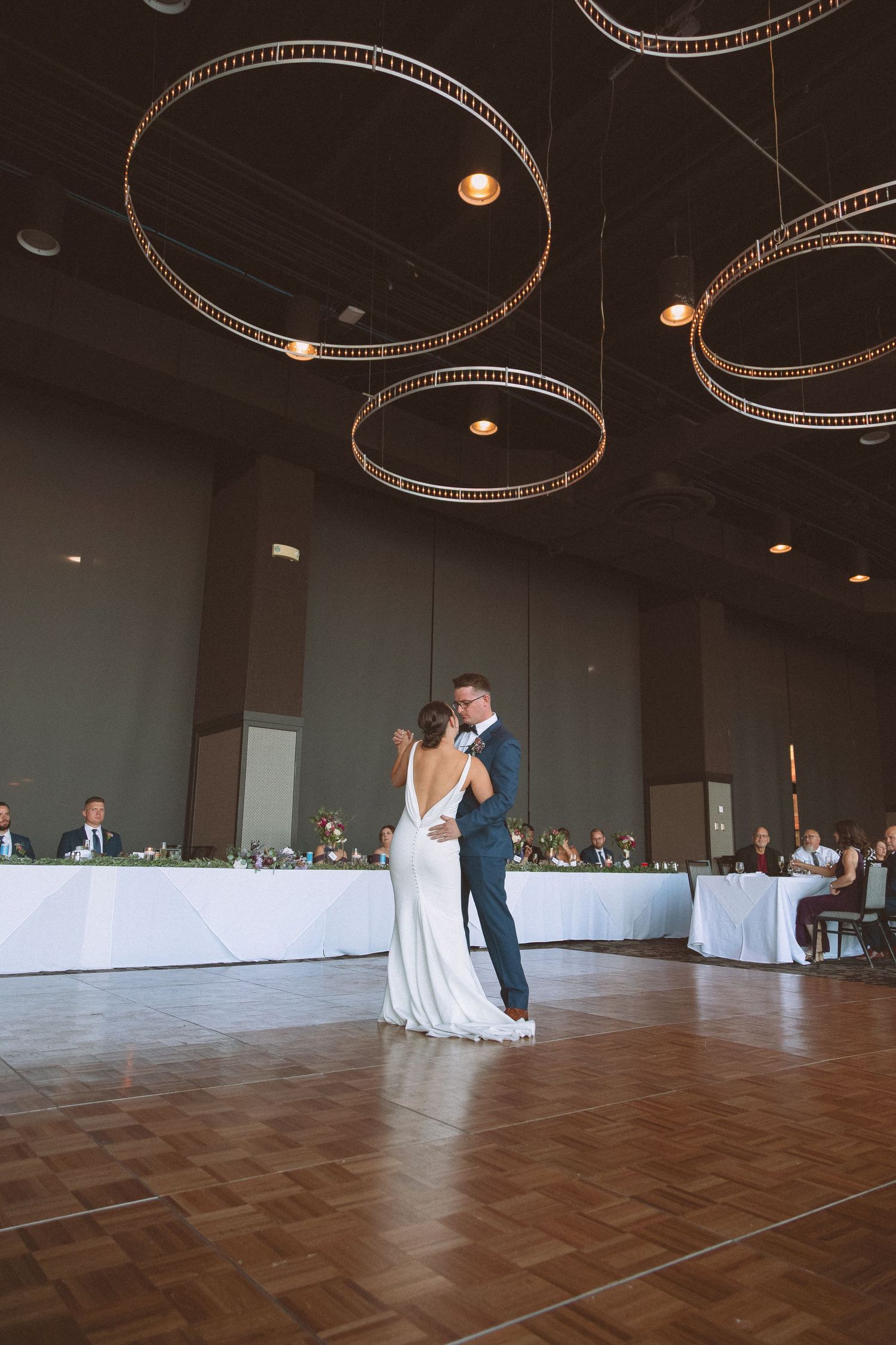 Bride and groom share elegant first dance under circular chandelier lights in modern ballroom venue.
