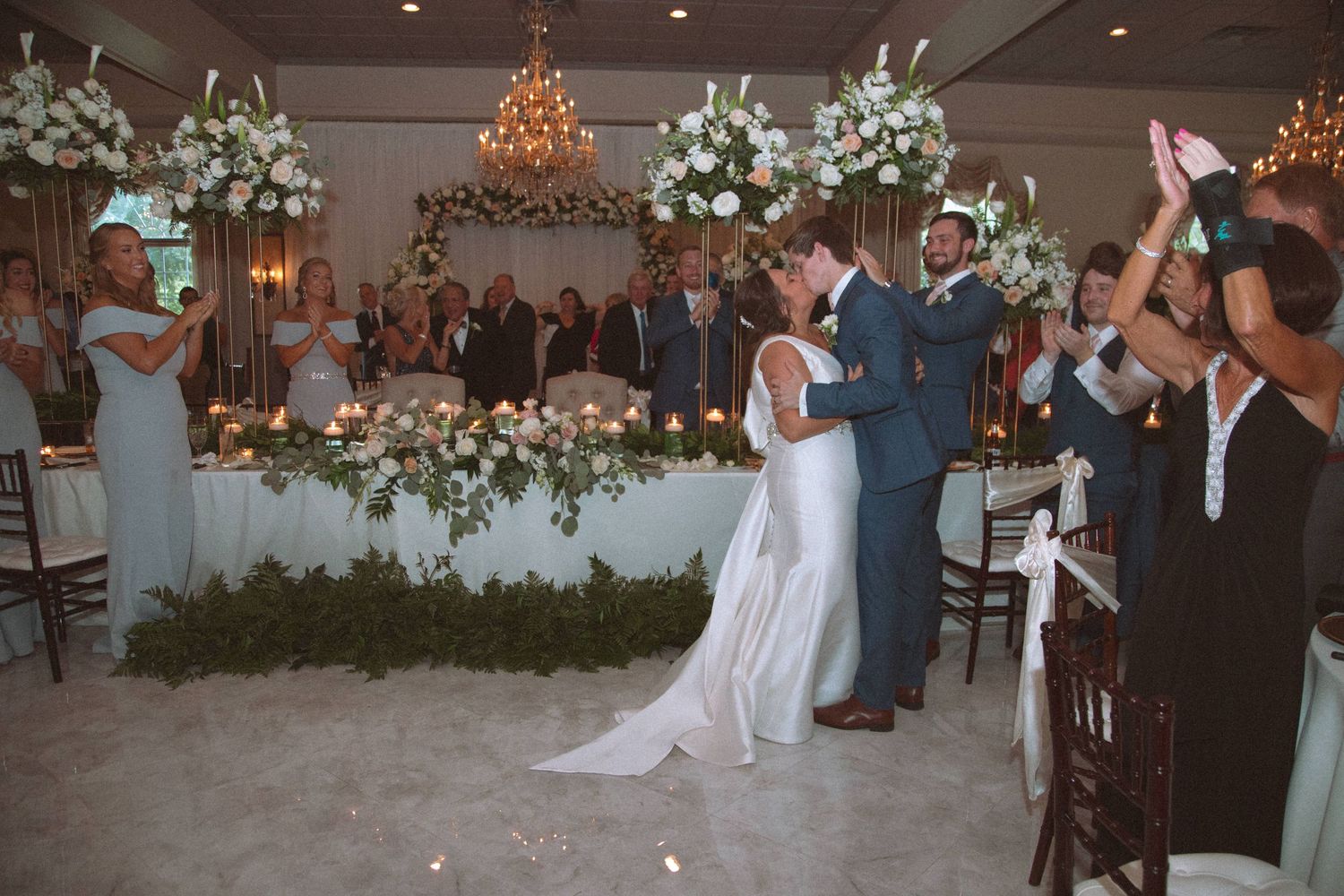 Bride and groom share first kiss at indoor wedding ceremony surrounded by white floral arrangements.