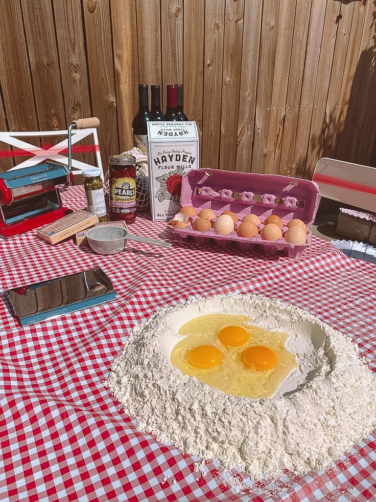 Pasta ingredients arranged on red checkered tablecloth with eggs in flour well, wine and hot sauce bottles.