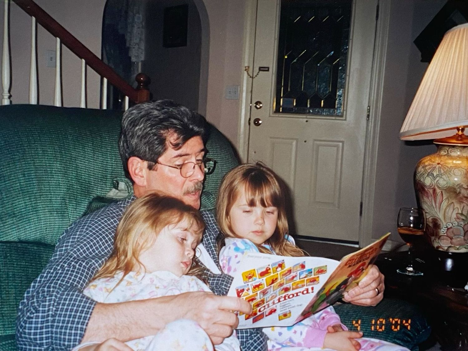 Grandfather reading a colorful picture book to two young girls while sitting in a green armchair at home.