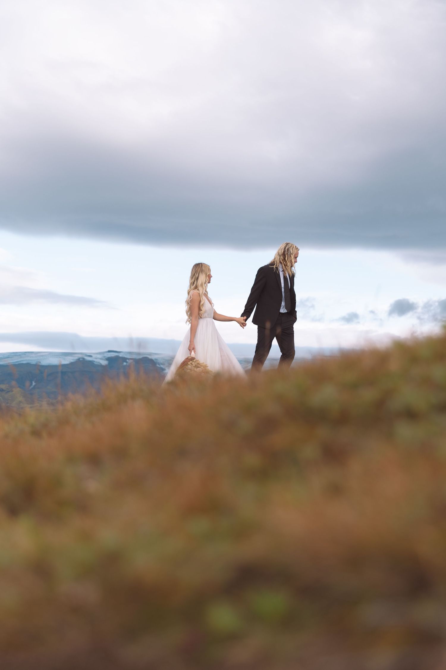 Bride and groom holding hands walking on grassy hillside under cloudy sky.
