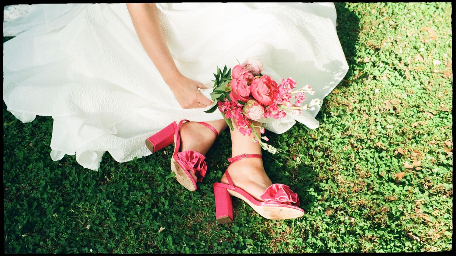 Bride holding pink peony bouquet wearing bold pink block heel sandals on green grass.