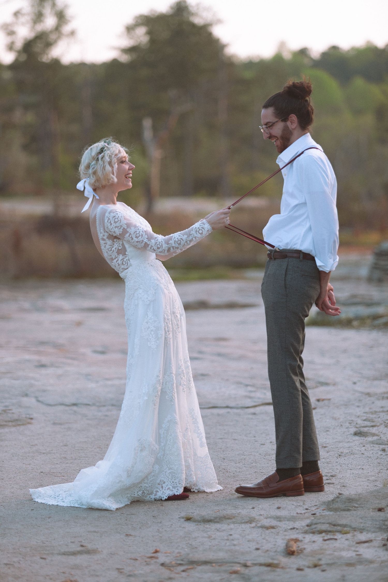 Bride in lace gown playfully holds groom's suspenders on a sandy outdoor wedding venue at dusk.