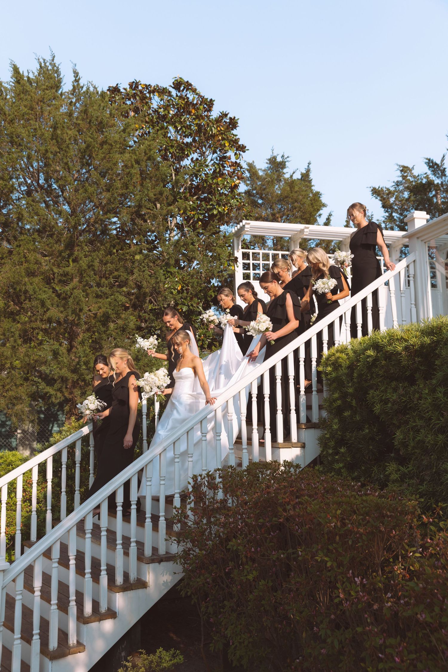 Bride in white gown descends grand staircase with bridesmaids in black dresses holding bouquets.