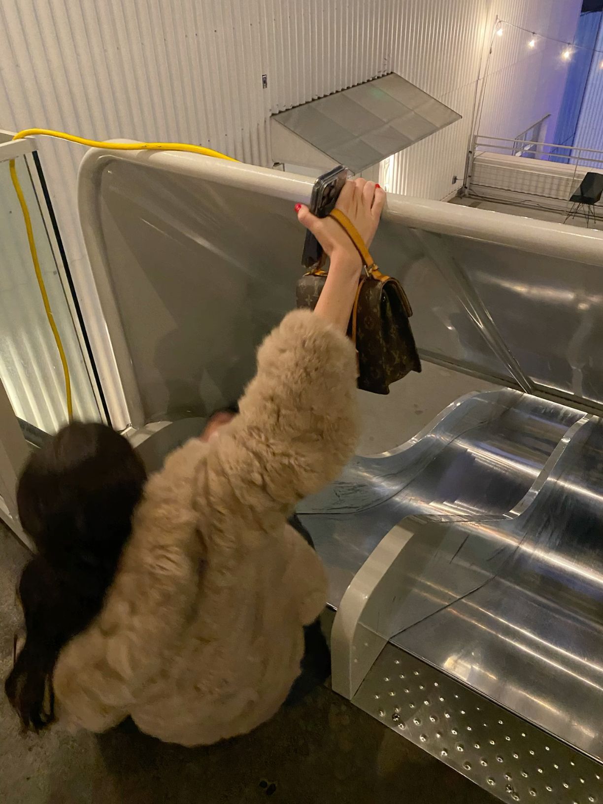 Woman in a fluffy coat reaching over an escalator railing holding a Louis Vuitton bag at night.
