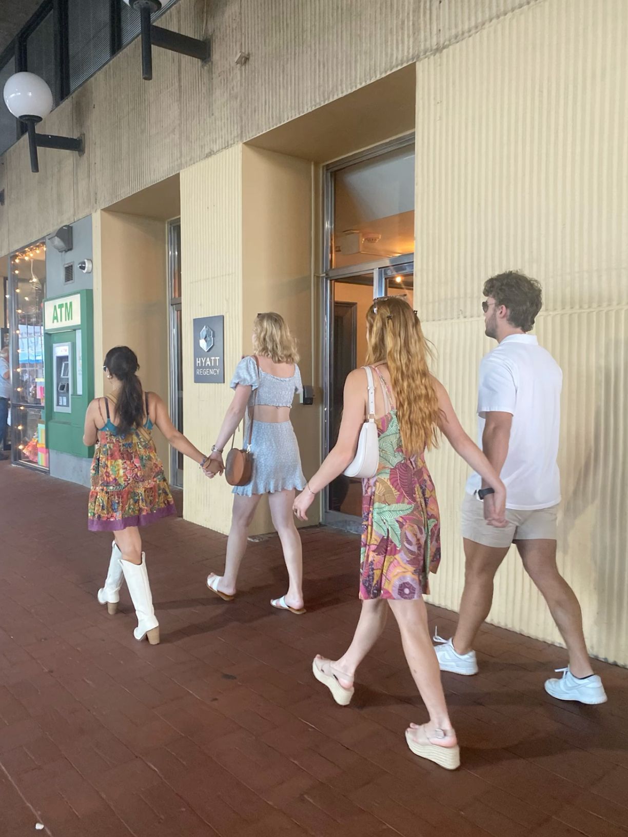 Group of four young adults in summer outfits walking toward entrance of a shopping center with ATM nearby.