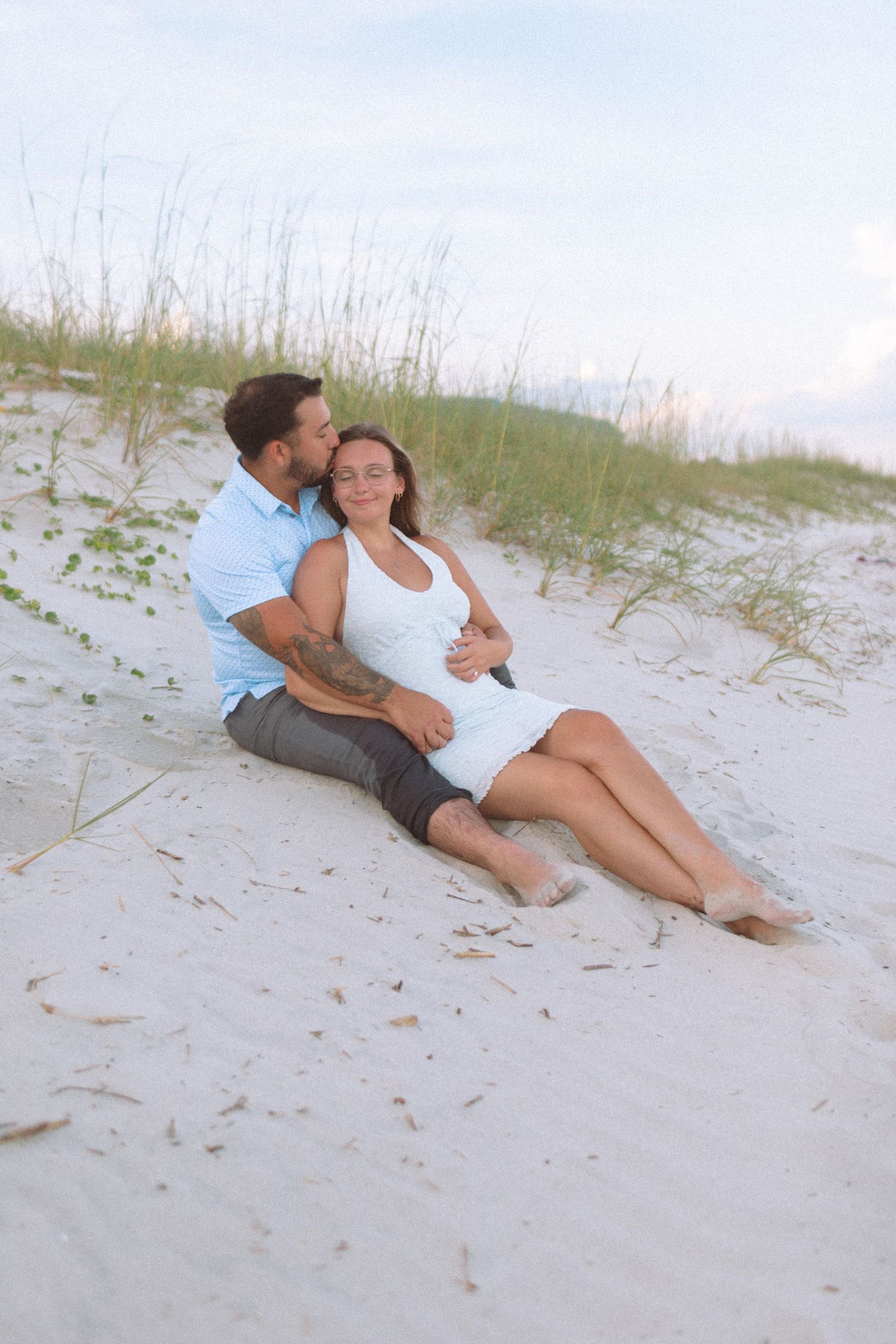 Couple relaxing together on white sand dunes, man embracing woman from behind near beach grass.