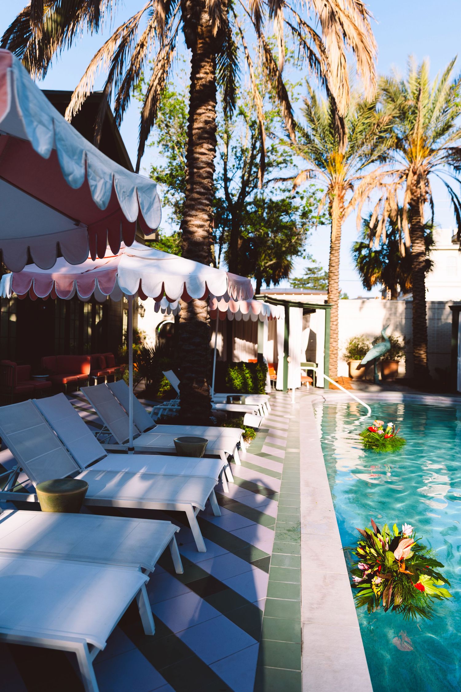 Luxury hotel pool with white lounge chairs, pink striped umbrella, tropical palm trees, and floral float on a sunny day.