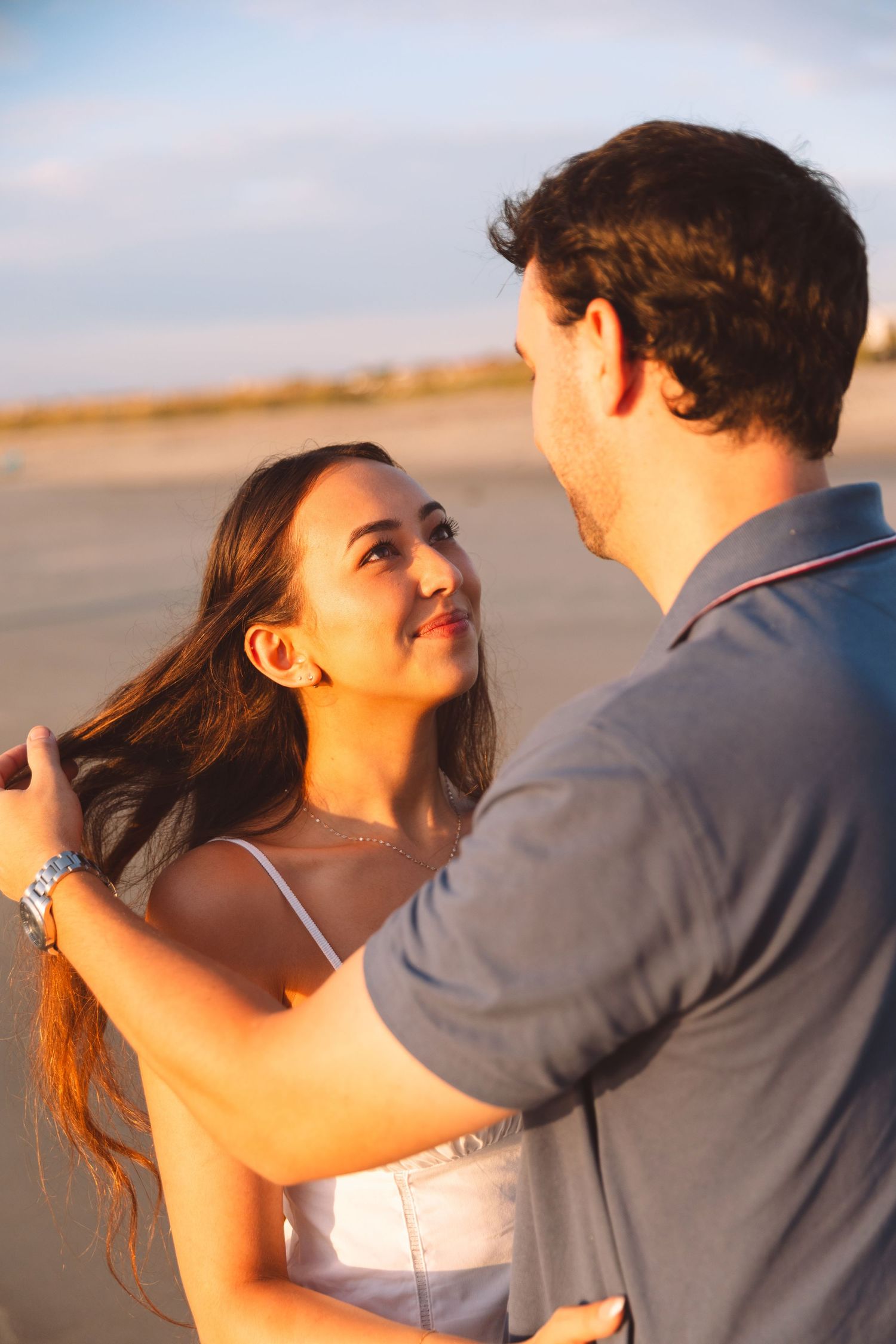 Couple sharing a romantic moment at sunset on a beach, woman smiling up at man holding her hair.