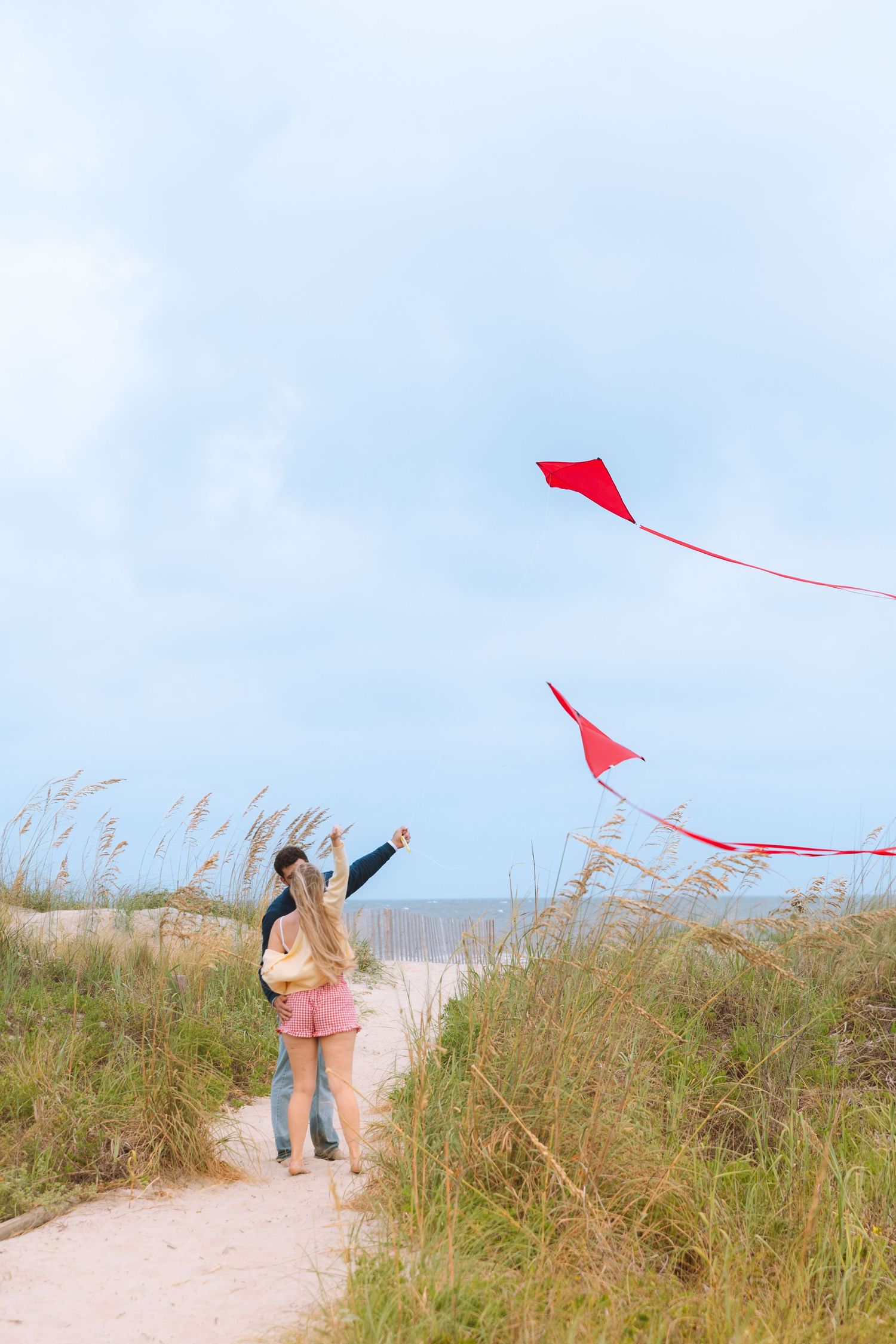 Couple flying a red kite on a sandy beach path surrounded by tall grass under a cloudy sky.