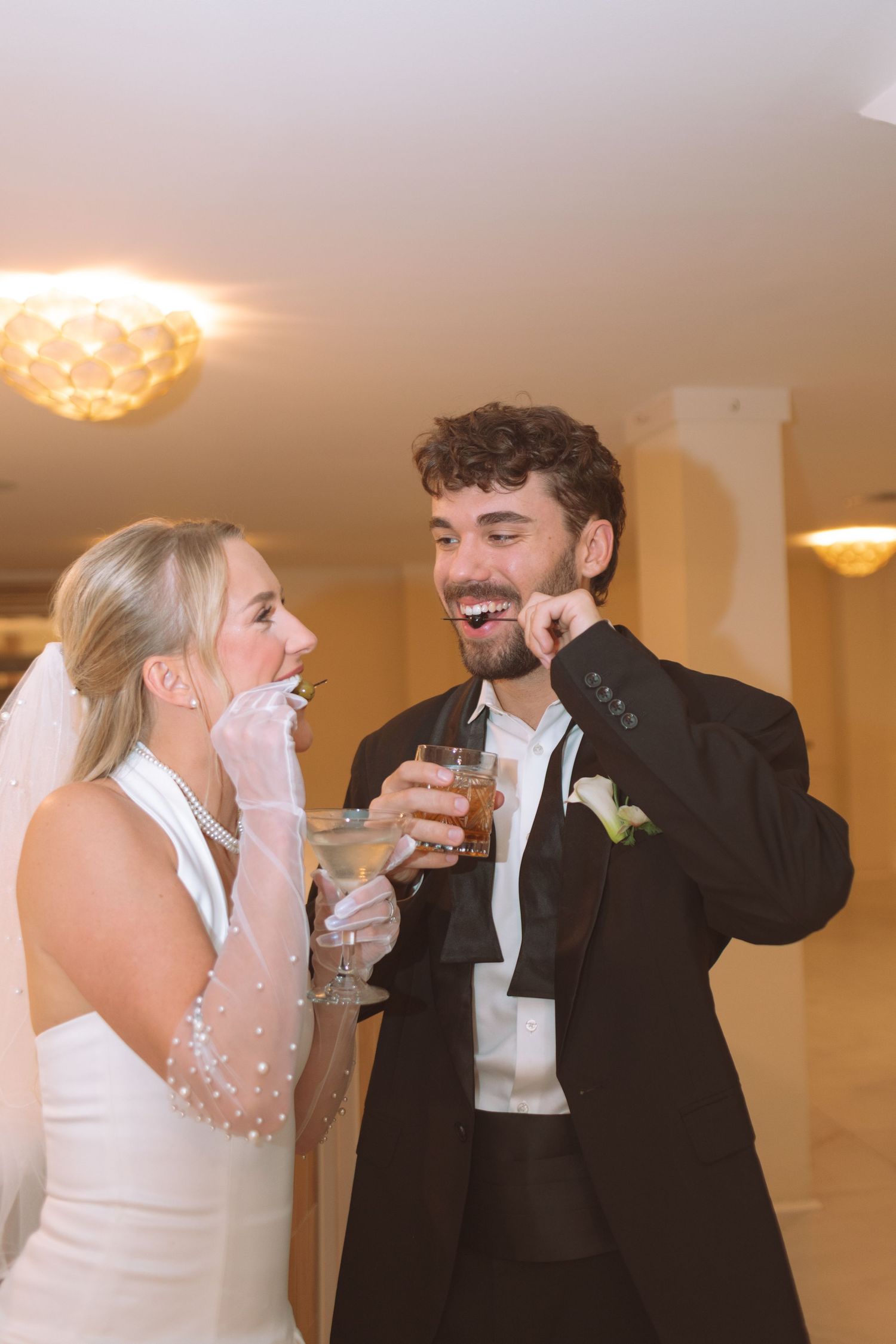 Laughing bride and groom clinking cocktail glasses at their wedding reception indoors.