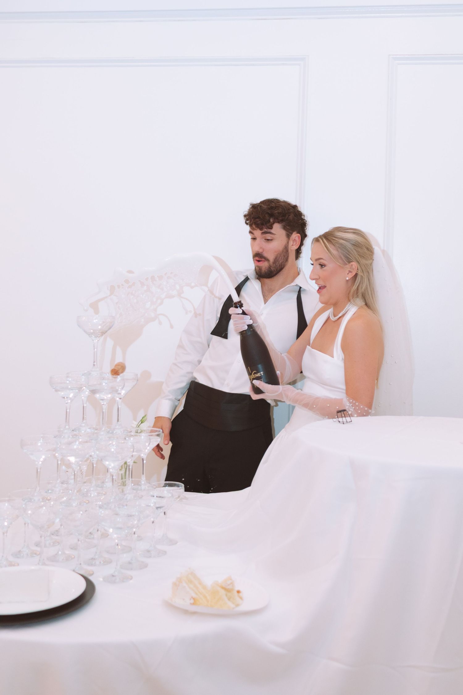Newlyweds excitedly pouring champagne into a tall coupe glass tower at their elegant wedding reception.